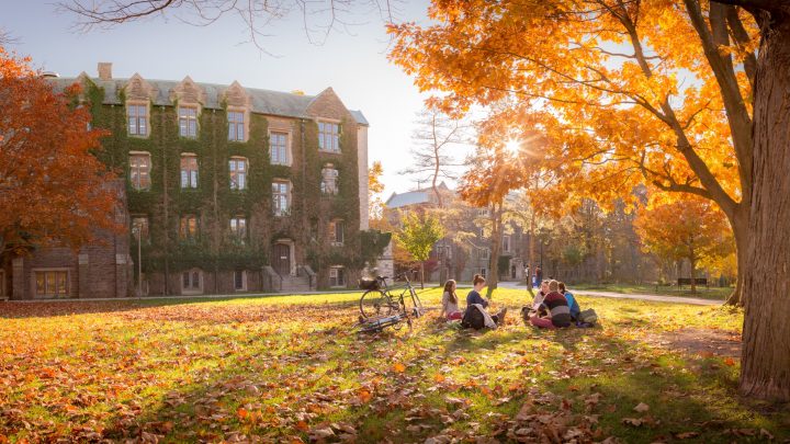 Area behind University Hall in the Fall, students sit under a tree.